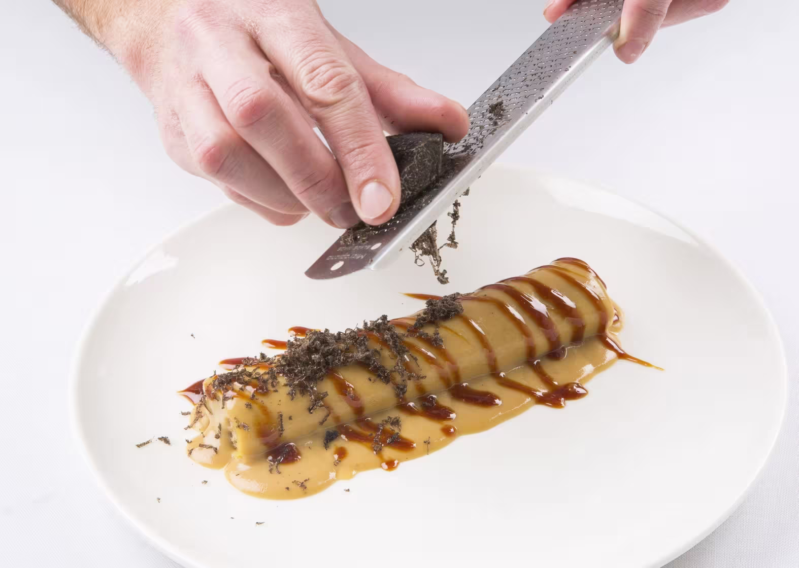 Plato de canelón bañado en salsa, decorado con trufa rallada al momento, servido sobre plato blanco en una presentación gourmet.