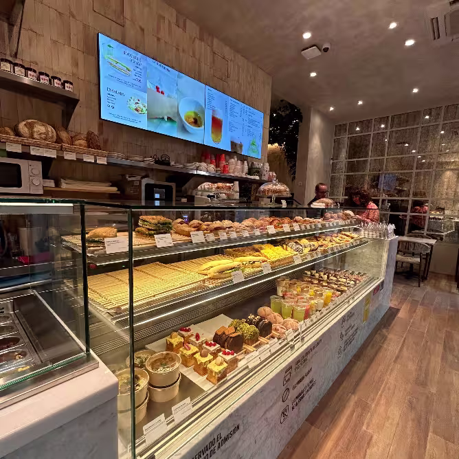 Vitrina iluminada con bollería, sándwiches, tartas y zumos bajo un menú digital en Maison Kayser Zurbano.