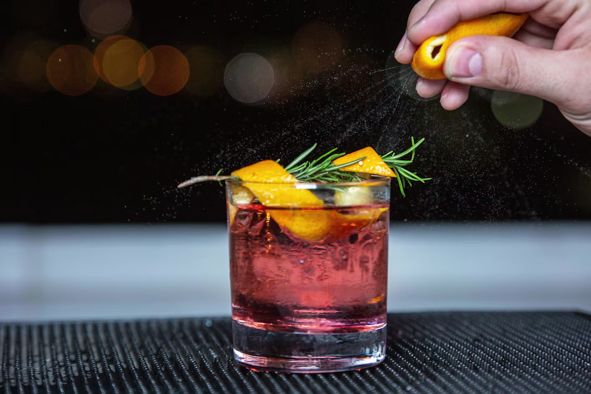 A hand squeezes an orange peel over a glass of red cocktail garnished with rosemary and orange peel on a dark surface.