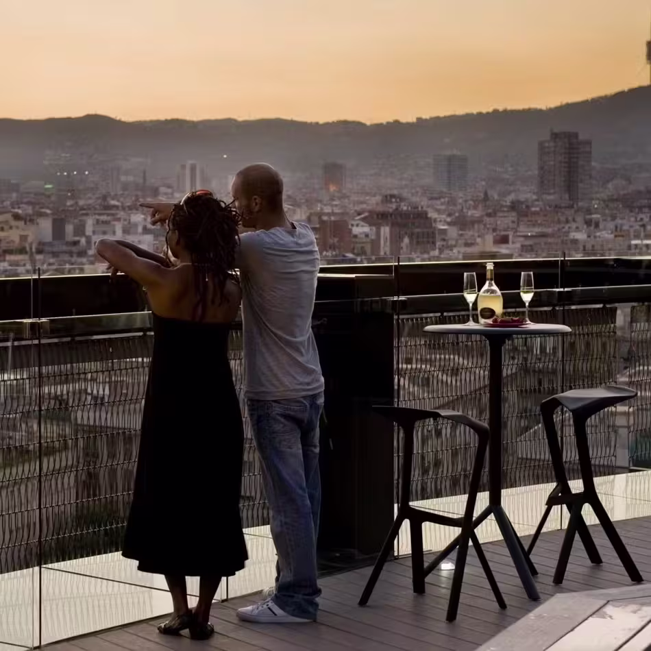 Dos personas de pie cerca una de la otra en la terraza de 360 Barceló Raval. Vista panorámica de la ciudad al fondo. La terraza tiene muebles modernos, incluyendo taburetes altos y una mesa con una elegante lámpara que refleja los colores del cielo.
