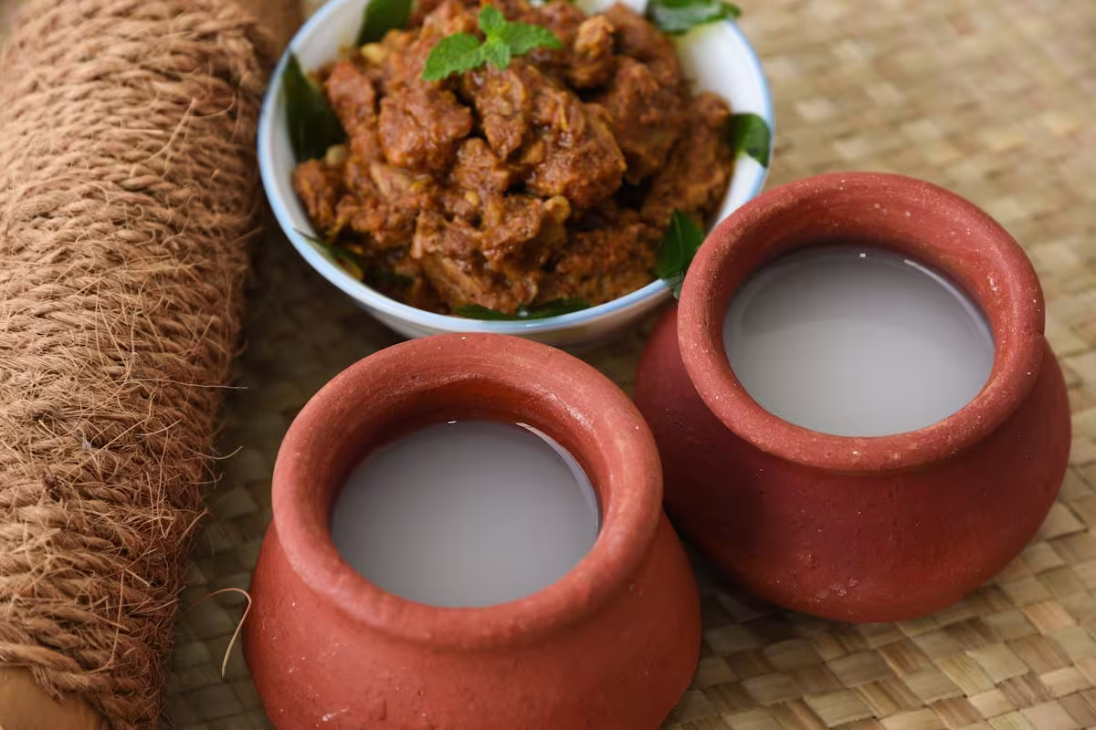 Two clay pots filled with a light-colored beverage are placed next to a bowl of spiced meat curry on a woven mat.