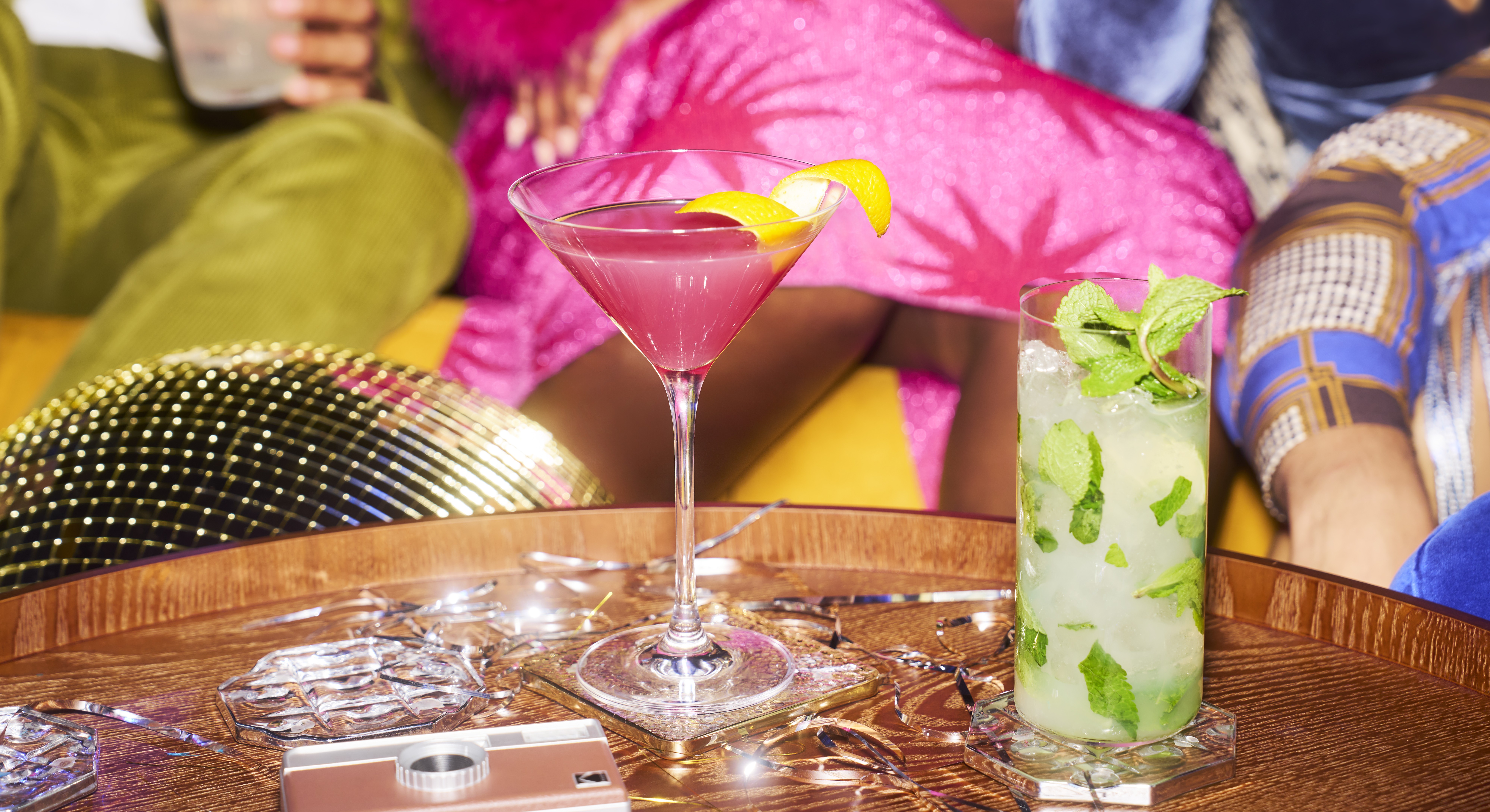 Two cocktails on a table in the foreground as partygoers celebrate in the background