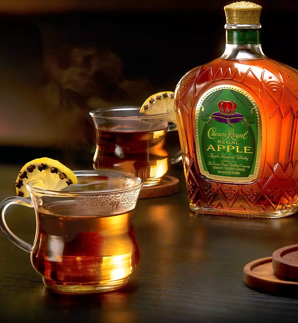 A bottle of Crown Royal Regal Apple whiskey beside two glass mugs of hot beverage garnished with clove-studded lemon slices.