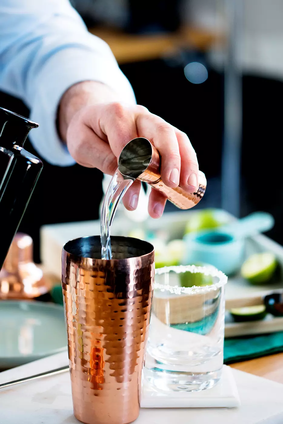 A person pours liquid from a jigger into a hammered copper cocktail shaker next to a glass with a salted rim and sliced limes in the background.