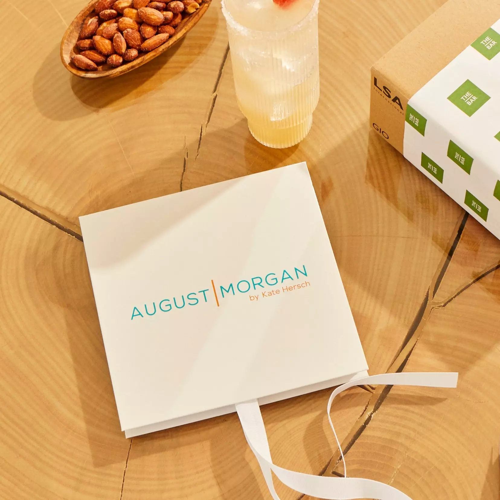 A square box labeled AUGUST MORGAN by Kate Hersch is on a wooden table next to a drink, a bowl of almonds, and part of a cardboard package.
