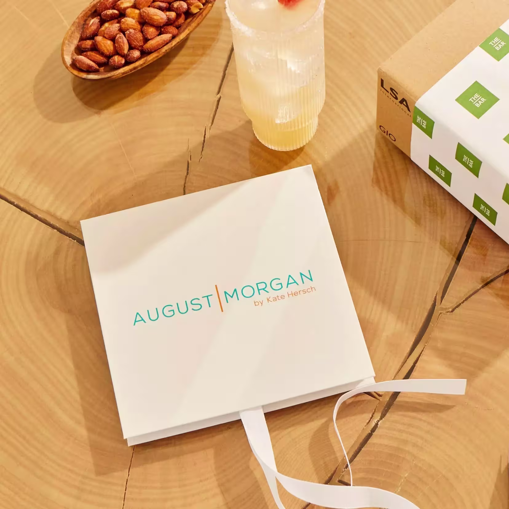 A square box labeled AUGUST MORGAN by Kate Hersch is on a wooden table next to a drink, a bowl of almonds, and part of a cardboard package.