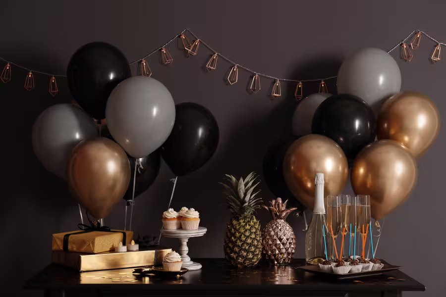 A table set for a celebration with cupcakes, pineapples, wrapped gifts, champagne, glasses, and bunches of black, gray, and gold balloons against a dark wall with string lights.