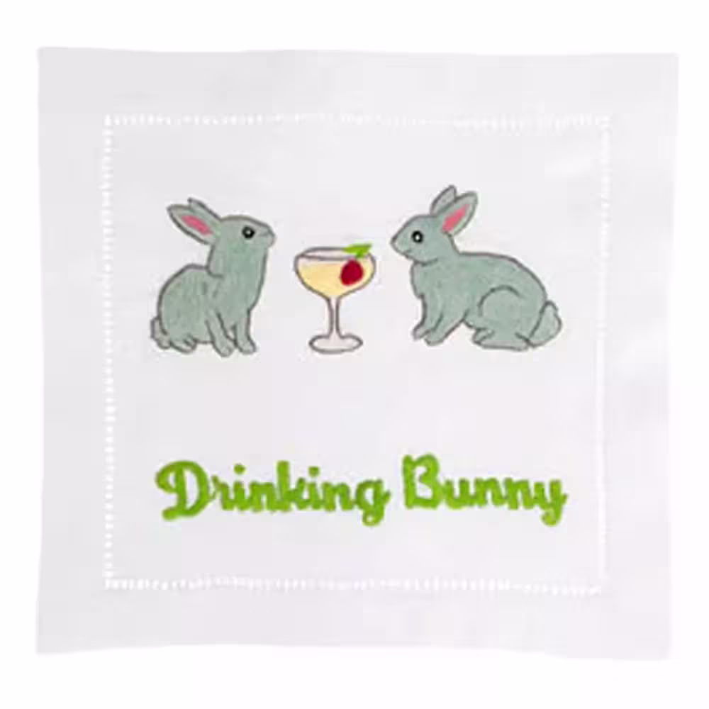 Two embroidered gray rabbits face a cocktail glass with an olive, above green text reading “Drinking Bunny” on a white cloth background.