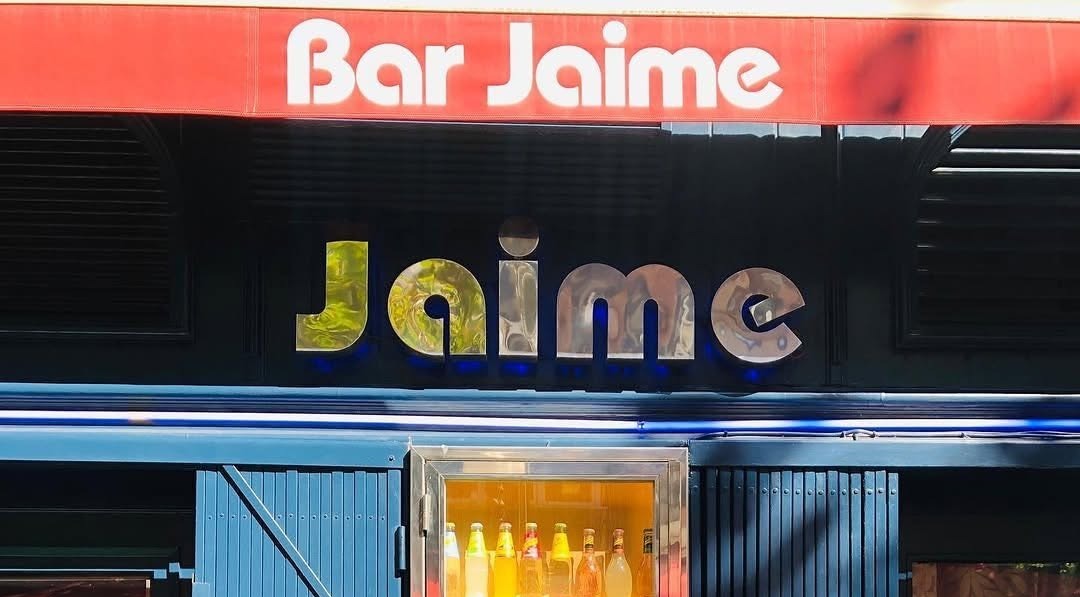La imagen muestra la fachada del Bar Jaime con su letrero rojo y azul. Destaca un frigorífico iluminado con botellas Schweppes en exhibición.