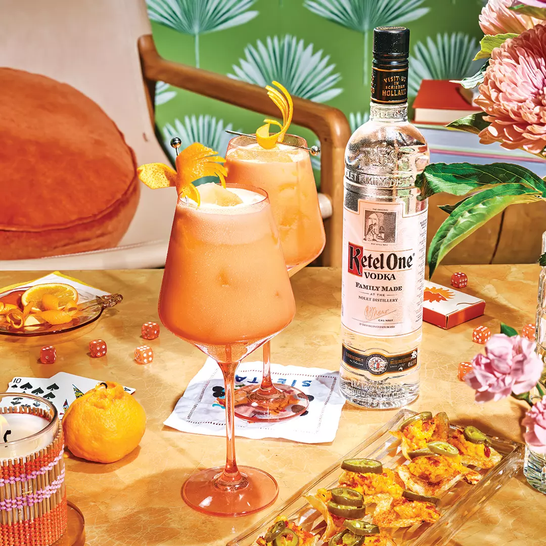 A bottle of Ketel One vodka and two orange cocktails on a table with snacks, playing cards, dice, and vibrant decor in the background.