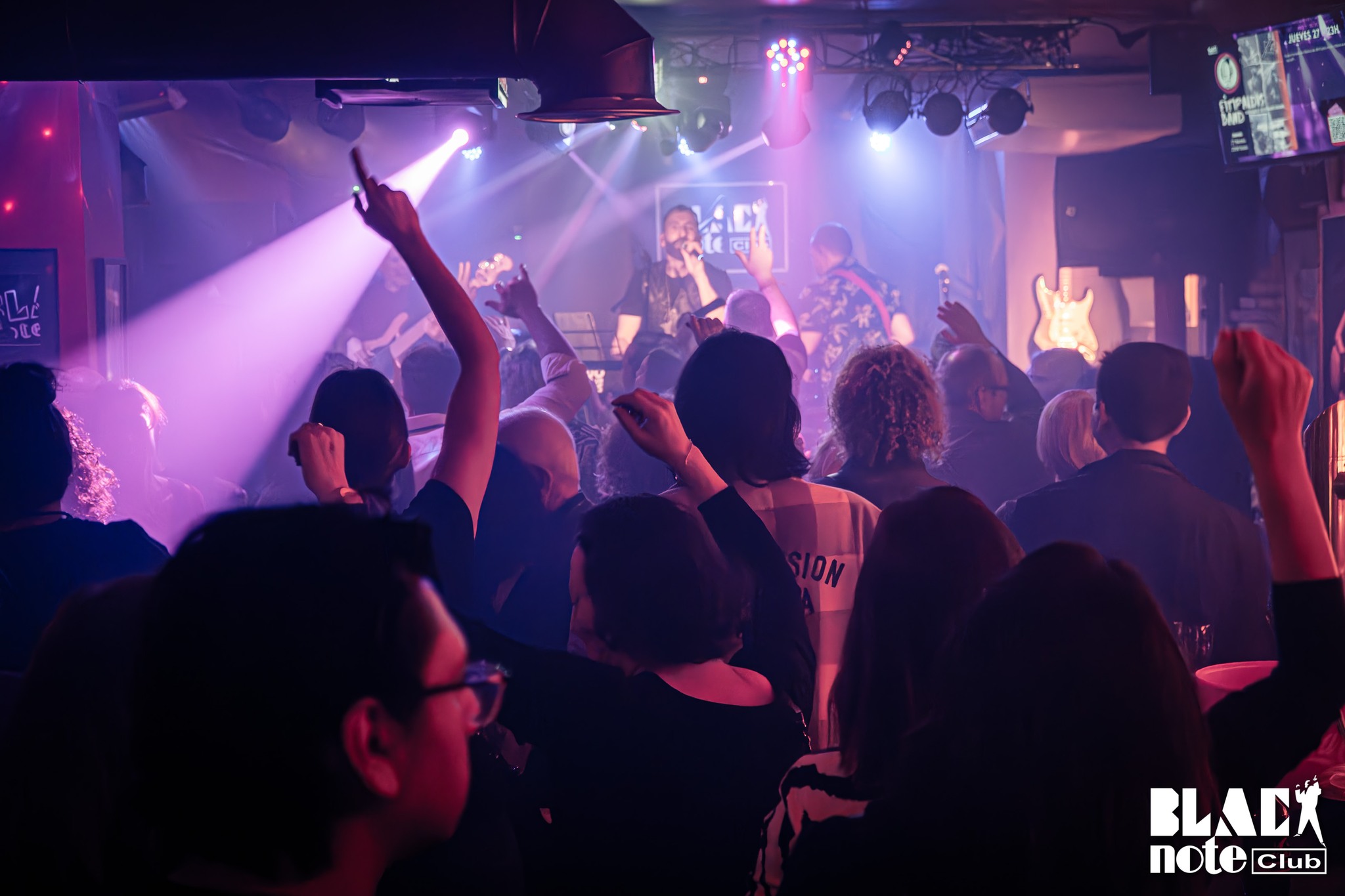 Concierto en directo en Black Note Club con luces violetas, público entregado y músicos sobre el escenario en un ambiente íntimo y vibrante de sala legendaria.