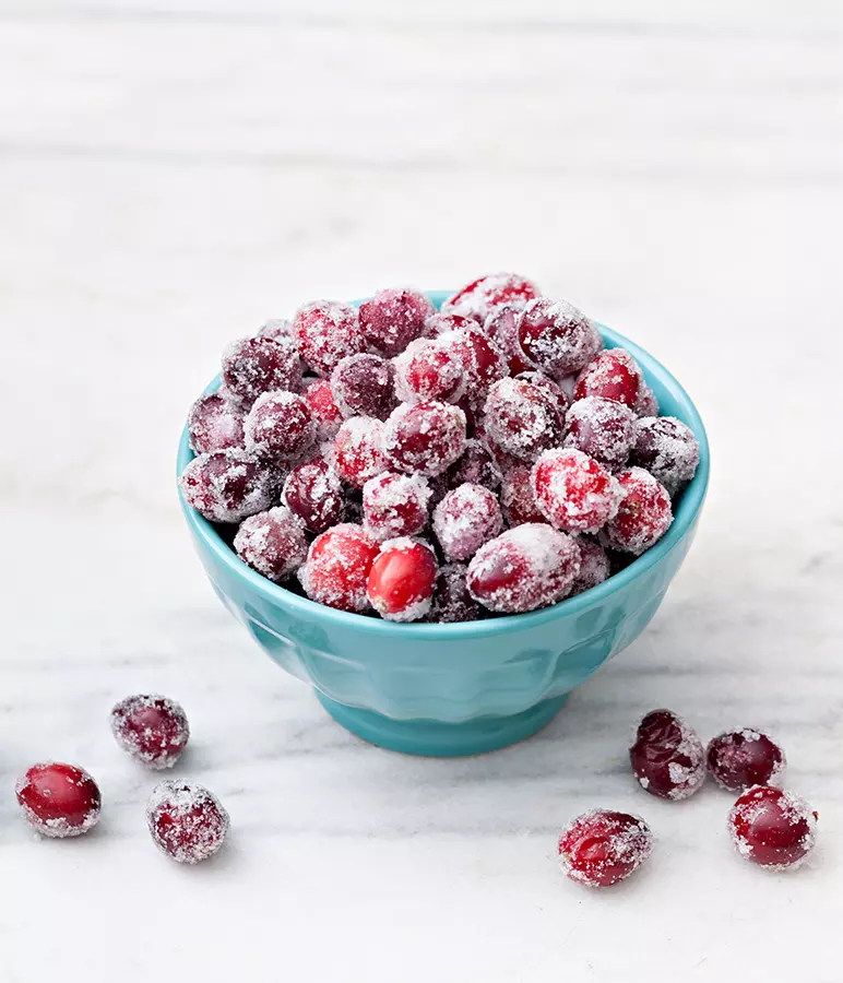 A blue bowl filled with sugared cranberries sits on a white surface, with a few sugared cranberries scattered beside the bowl.