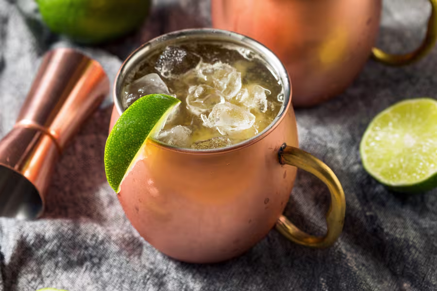 A copper mug filled with ice and a light-colored drink, garnished with a lime wedge, sits on a cloth next to cut limes and a metal jigger.