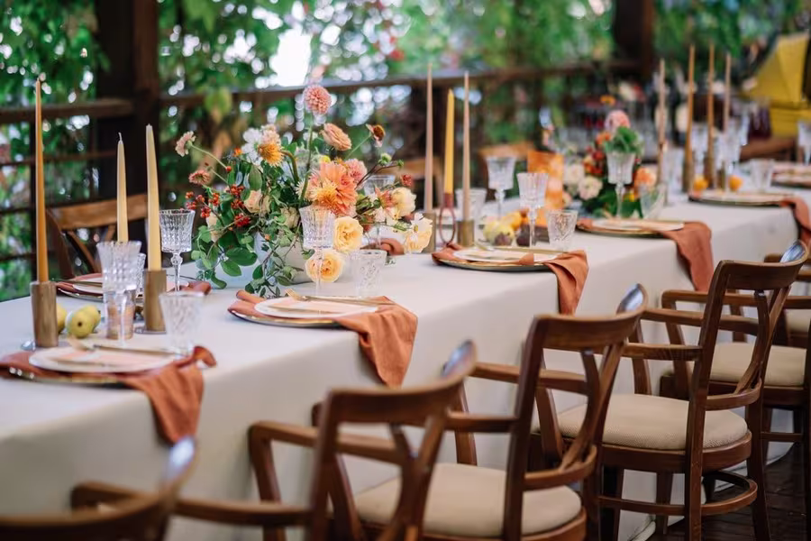 A long dining table is set with white tablecloth, orange napkins, glassware, plates, floral centerpieces, and tall candles, surrounded by wooden chairs in an outdoor setting.