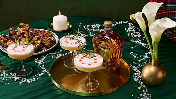 Three cocktails garnished with rosemary and red drops sit on a gold tray with a bottle of Crown Royal, a plate of dessert bars, a lit candle, and white calla lilies on a green tablecloth.