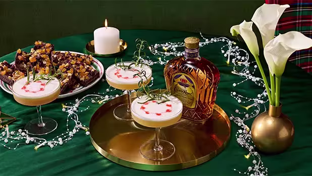 Three cocktails garnished with rosemary and red drops sit on a gold tray with a bottle of Crown Royal, a plate of dessert bars, a lit candle, and white calla lilies on a green tablecloth.