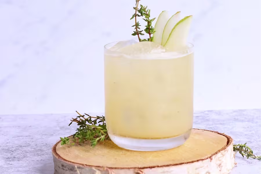 A cocktail garnished with pear slices and a sprig of thyme, served in a short glass with ice, placed on a wooden slab.