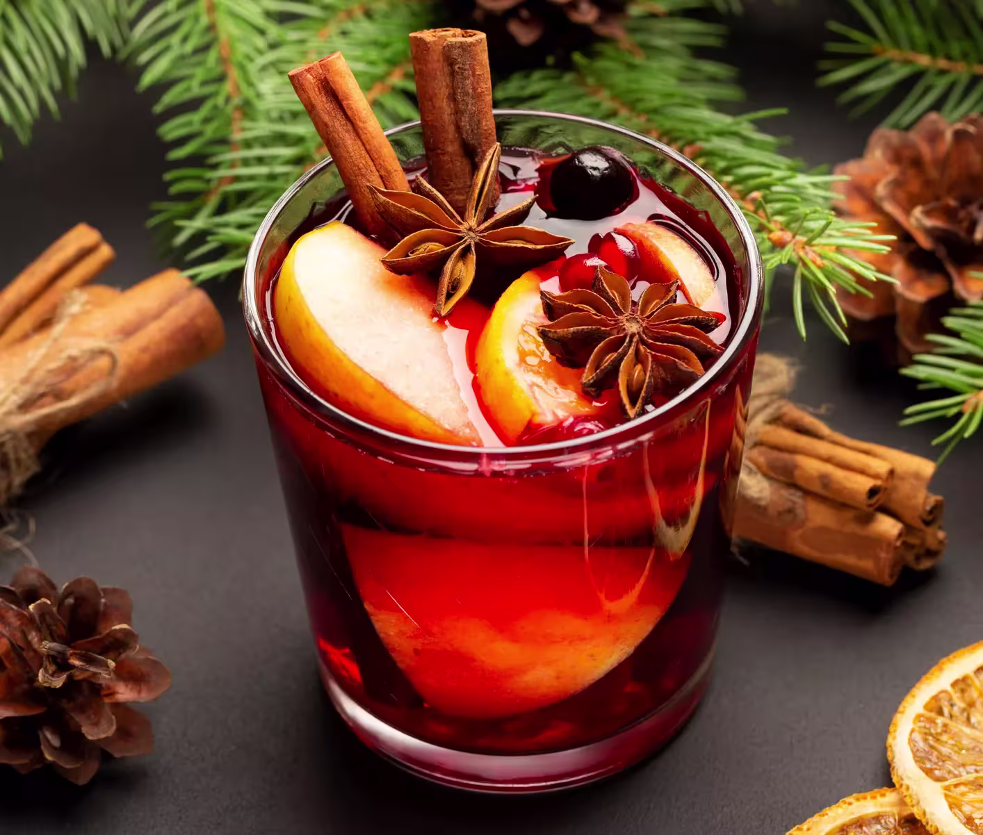 A glass of mulled wine garnished with apple slices, star anise, cinnamon sticks, and a cherry, with pine branches and pine cones in the background.