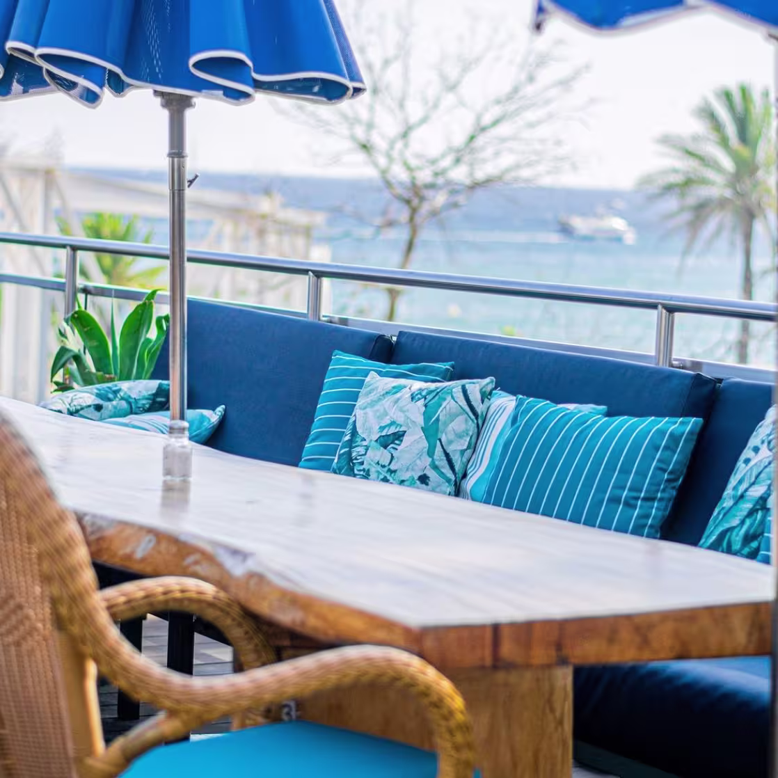 La imagen muestra una zona de asientos de Marina Bay al aire libre con vista a un cuerpo de agua. Hay sombrillas azules, una mesa de madera y sillas con cojines azules que tienen patrones de rayas. El entorno es un balcón