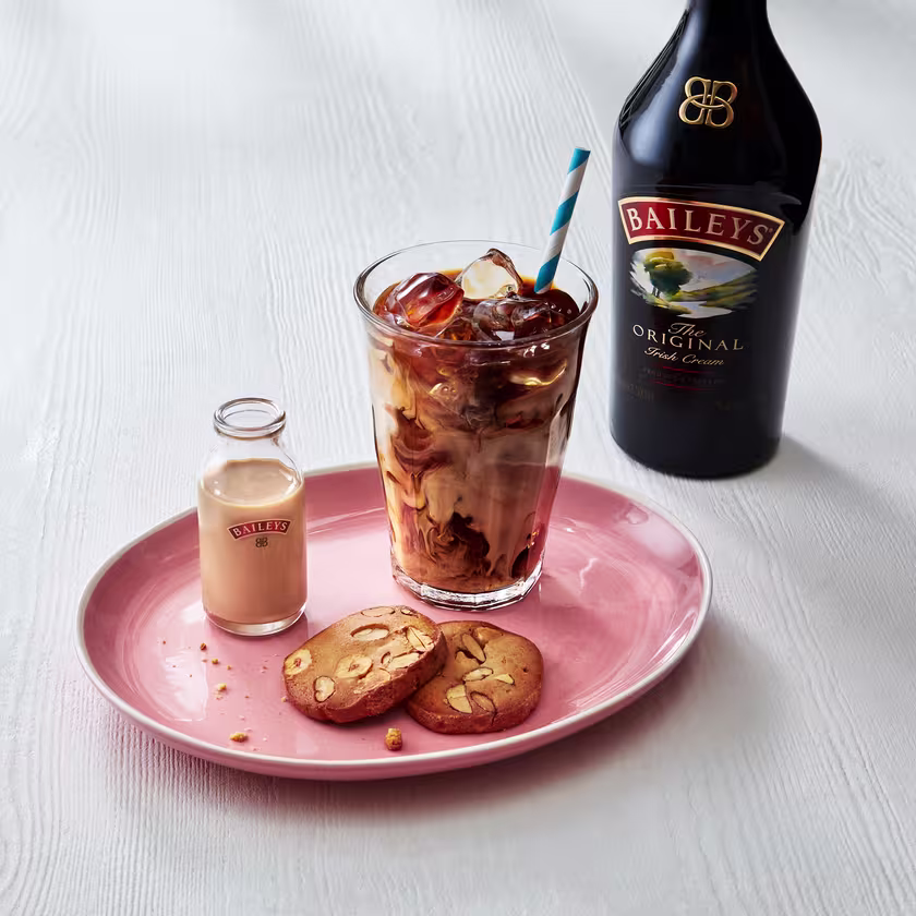 La imagen muestra una botella de Baileys Original Irish Cream, un plato con dos galletitas, una botella pequeña de Baileys y un vaso con el cóctel Frappé Helado con Baileys.
