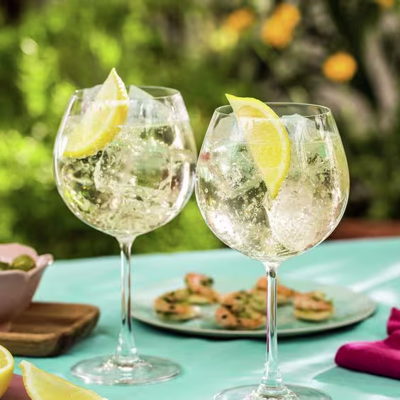 En la imagen se muestran dos copas de Gin and Tonic, adornadas con rodajas de limón. De fondo, una variedad de comidas.
