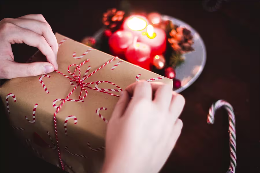 Hands tying red and white string around a gift wrapped in candy cane paper, with a lit candle and festive decorations in the background.