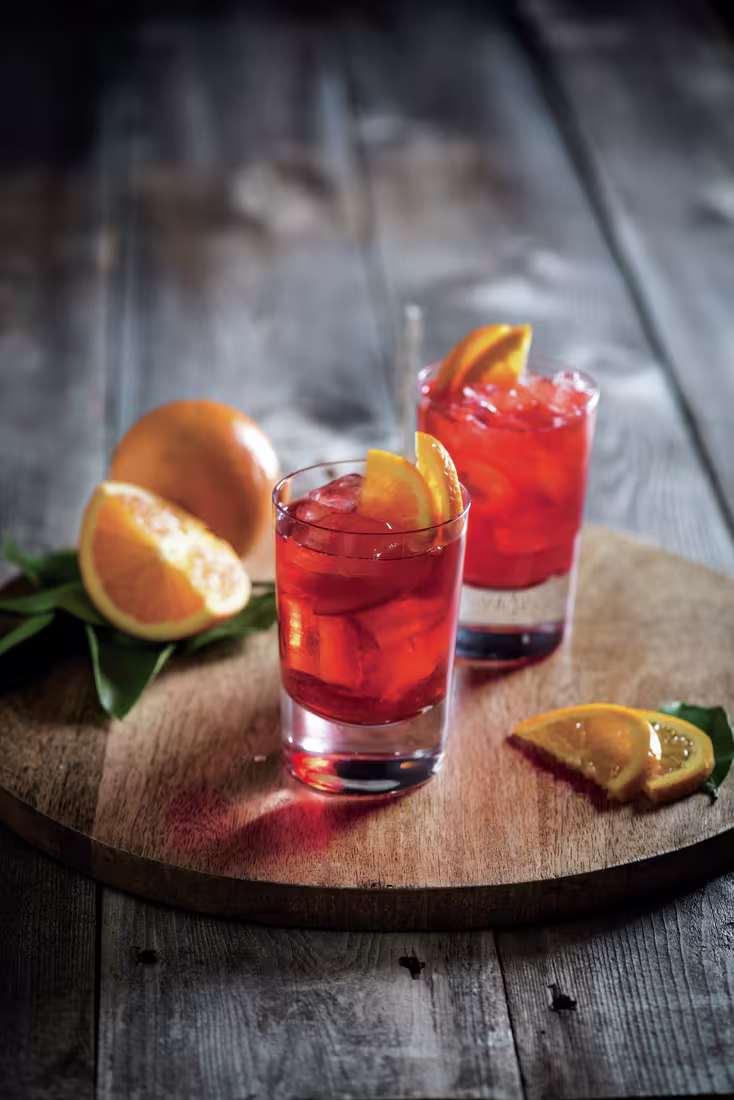 La imagen muestra dos vasos con el cóctel Negroni y varias rodajas de naranja alrededor.