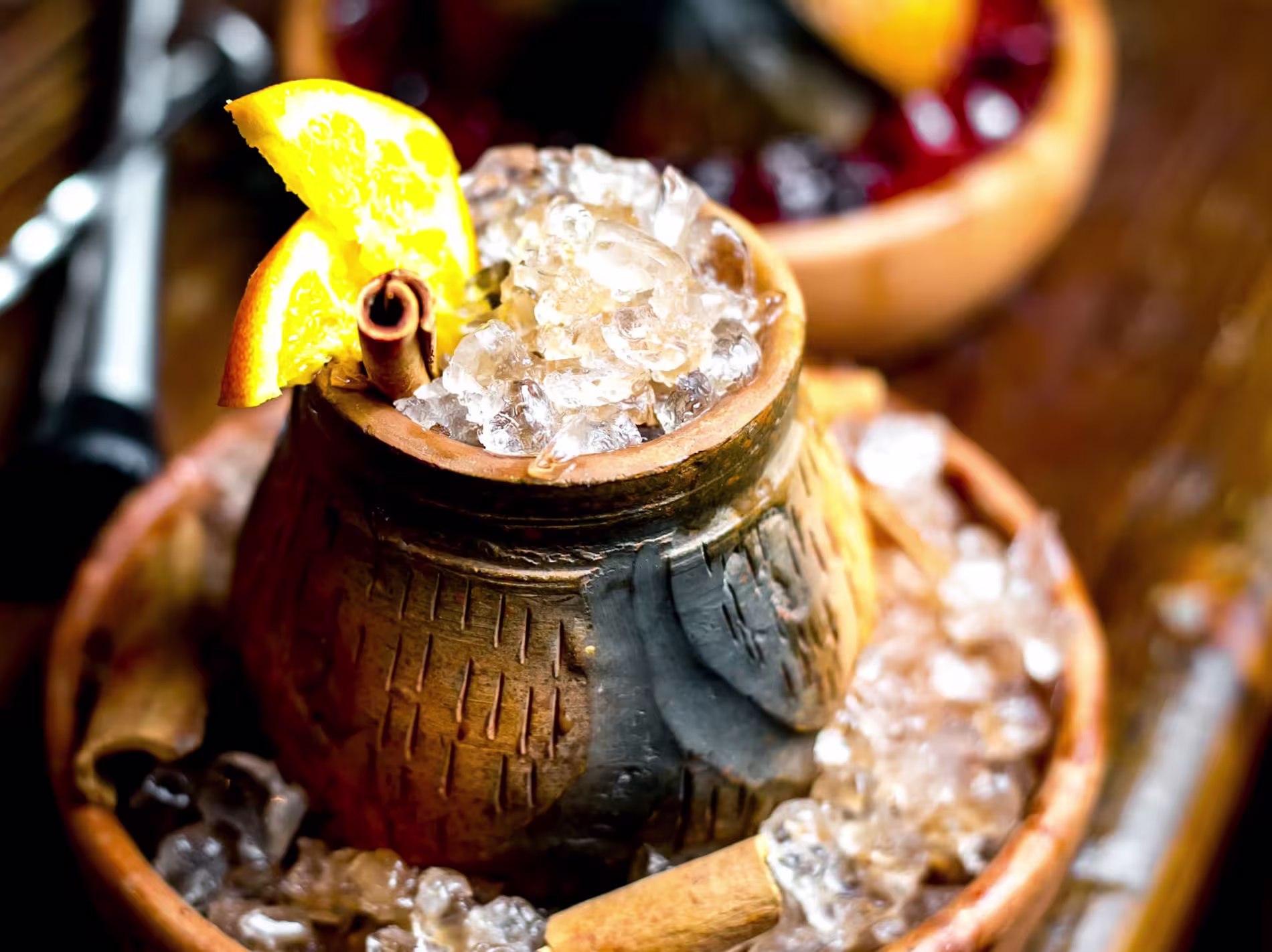 A rustic clay cup filled with crushed ice, garnished with orange slices and a cinnamon stick, sits in a larger bowl of ice on a wooden surface.