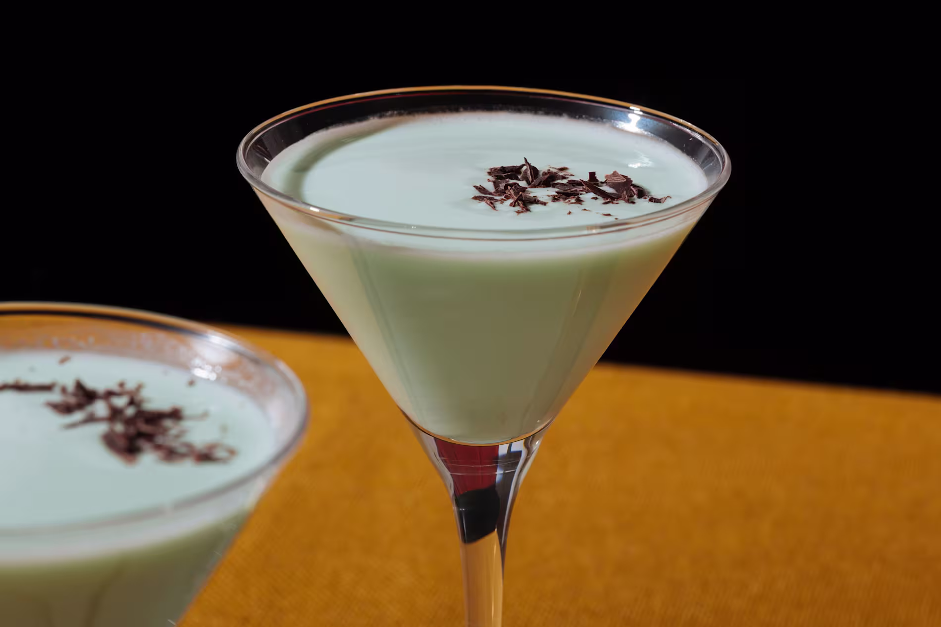 A pale green creamy cocktail in a martini glass, garnished with chocolate shavings, sits on a yellow surface with a black background.