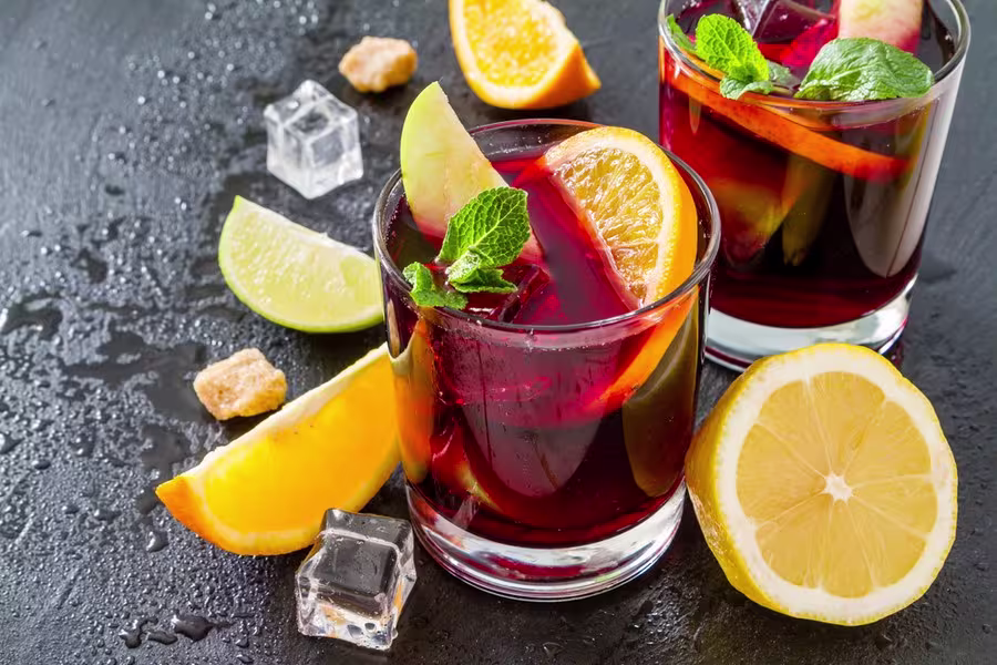 Two glasses of red sangria with ice, orange slices, apple slices, lemon, lime wedges, mint, sugar cubes, and ice cubes on a dark textured surface.