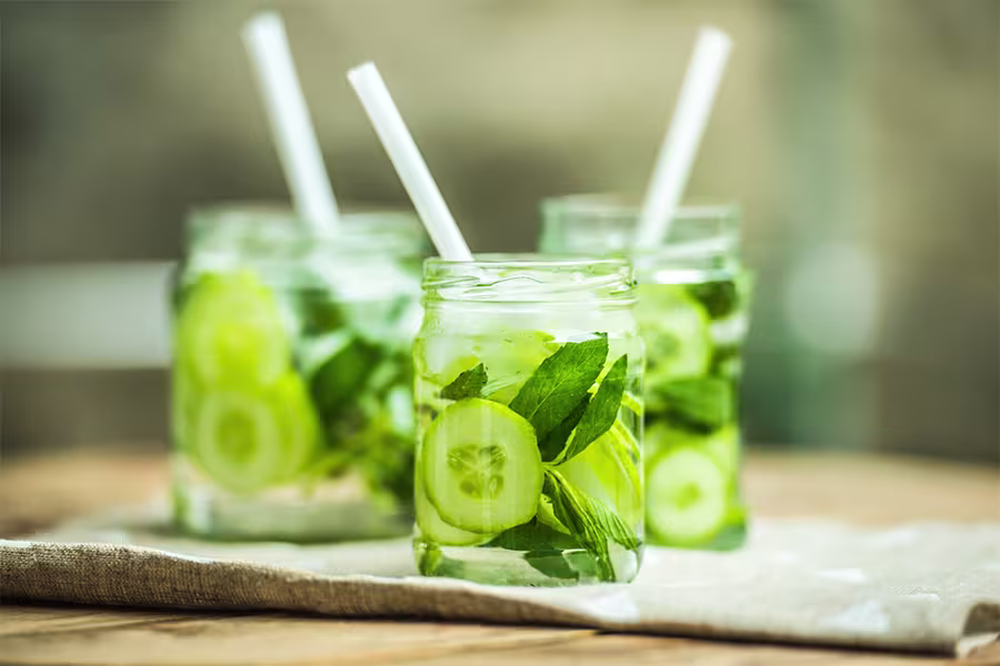 Three glass jars filled with cucumber slices, mint leaves, ice, and water, each with a white straw, sit on a beige cloth on a wooden surface.