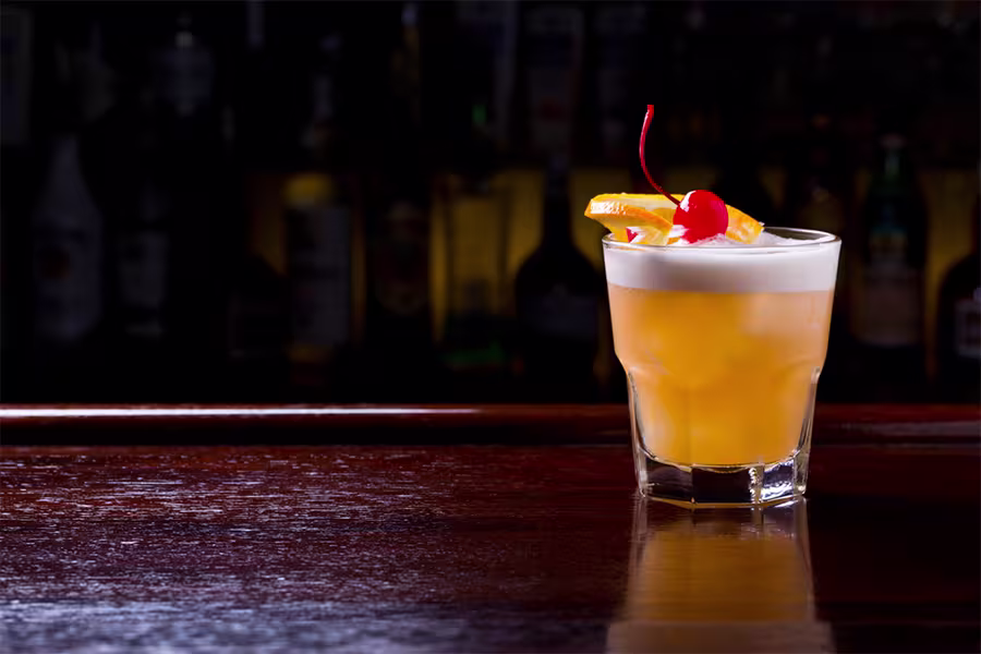 A whiskey sour cocktail garnished with an orange slice and a cherry sits on a polished wooden bar with a dark, blurred background.