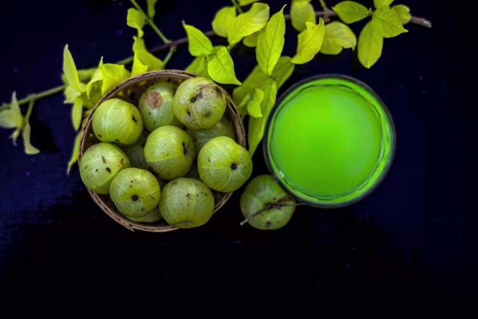 A basket of Indian gooseberries beside a glass of green juice and a branch with yellow-green leaves on a dark surface.