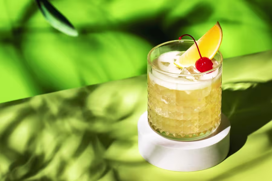 A glass of iced cocktail garnished with an orange slice and a cherry sits on a white coaster against a bright green background.
