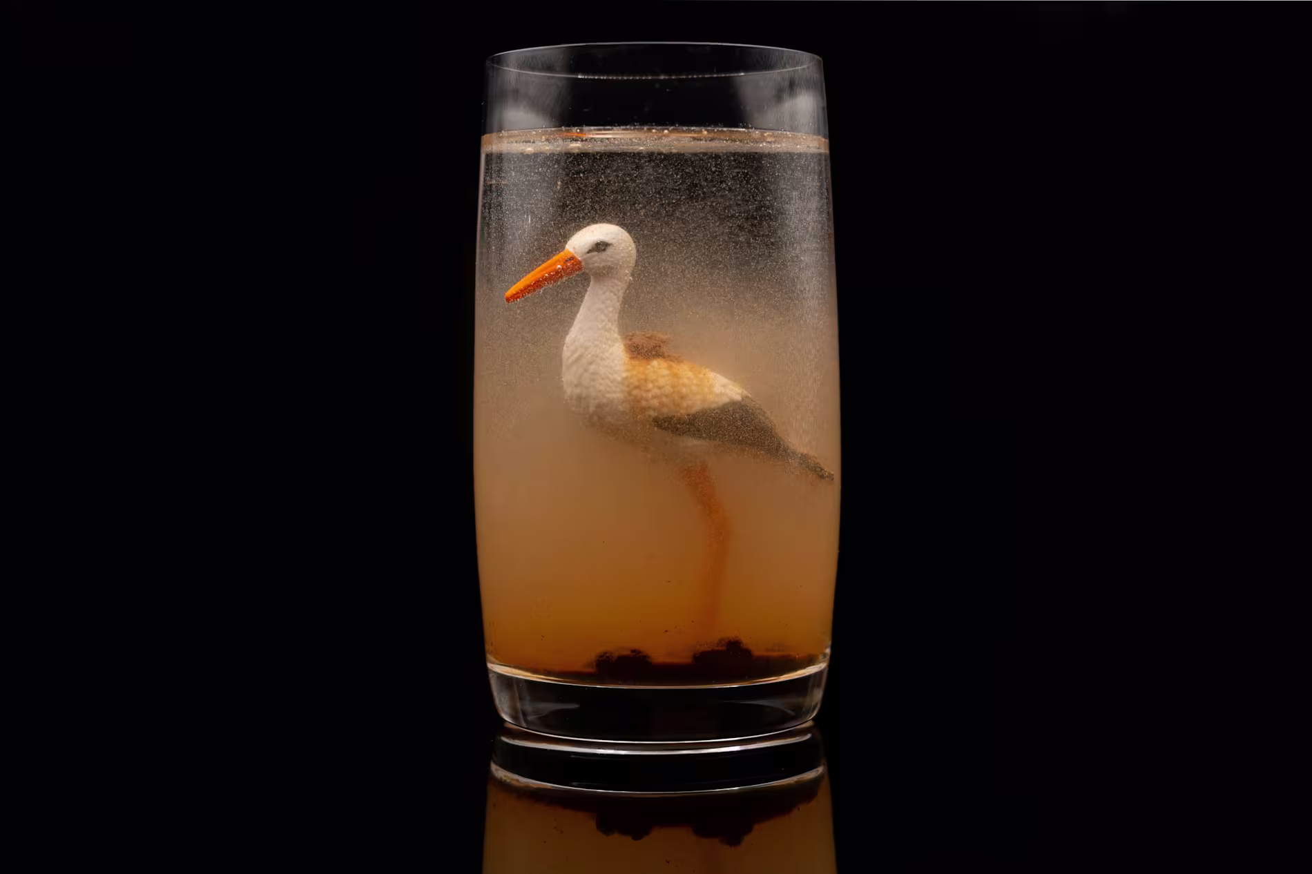 A toy stork figurine is submerged in a cloudy liquid inside a clear glass against a black background.