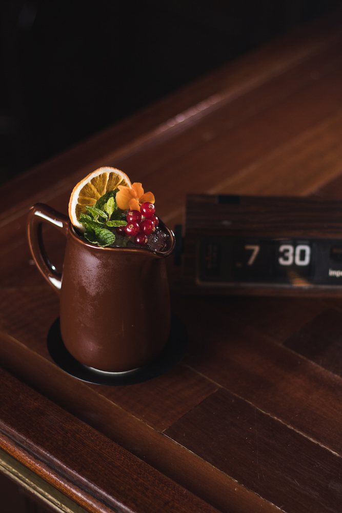 En Bobby's, un cóctel servido en una jarra de cobre se adorna con frutas frescas, menta y flores, sobre una elegante barra de madera.