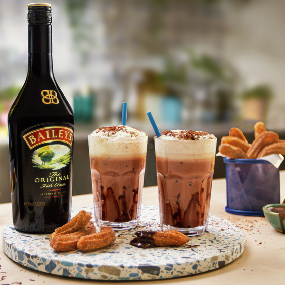 La imagen muestra tres vasos con el postre Baileys Salted Caramel Cheesecake, mientras que en el fondo se puede observar una botella de Baileys Salted Caramel.