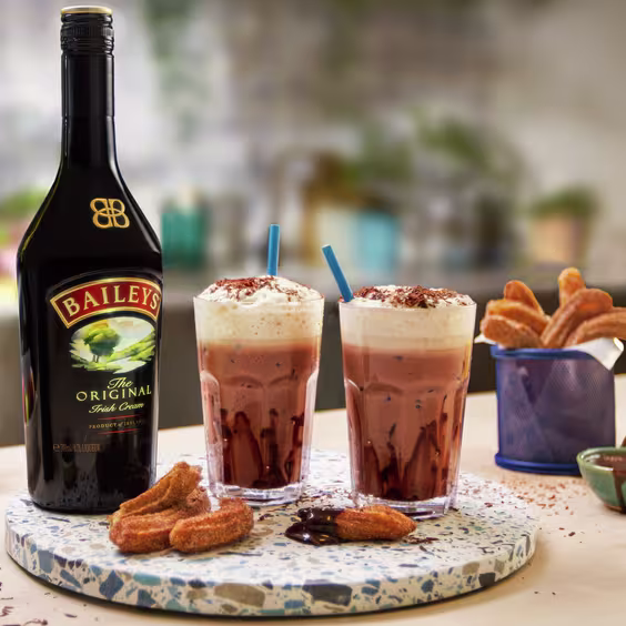 La imagen muestra tres vasos con el postre Baileys Salted Caramel Cheesecake, mientras que en el fondo se puede observar una botella de Baileys Salted Caramel.