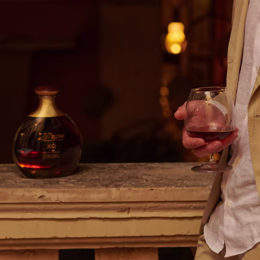 Una persona con traje beige sostiene un vaso de licor marrón, junto a una botella de Zacapa XO en una repisa de piedra.