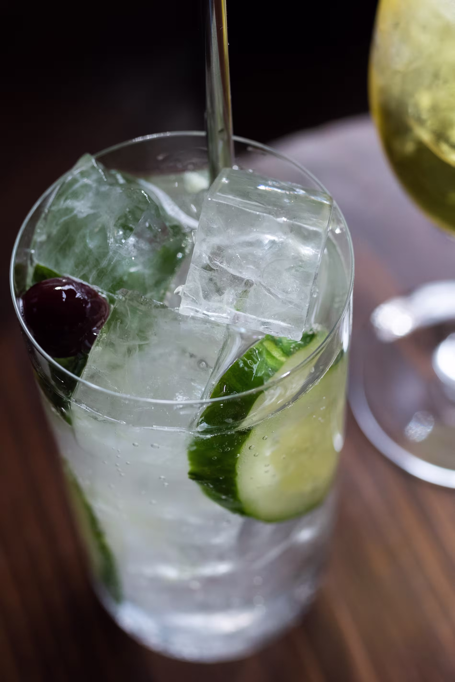 A clear glass filled with ice cubes, cucumber slices, and a dark olive, containing a clear liquid with a metal stirrer, sits on a wooden surface next to another drink.