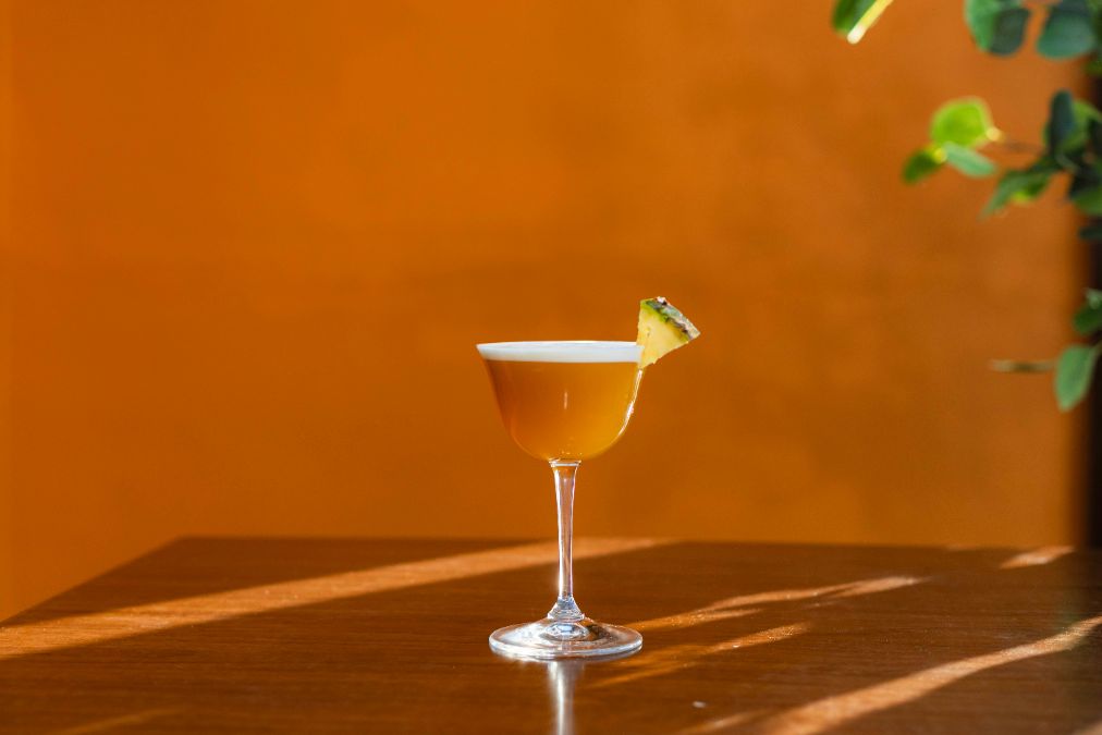 A cocktail with a frothy top and a pineapple garnish sits on a wooden table against an orange background, with sunlight streaming in from the side.