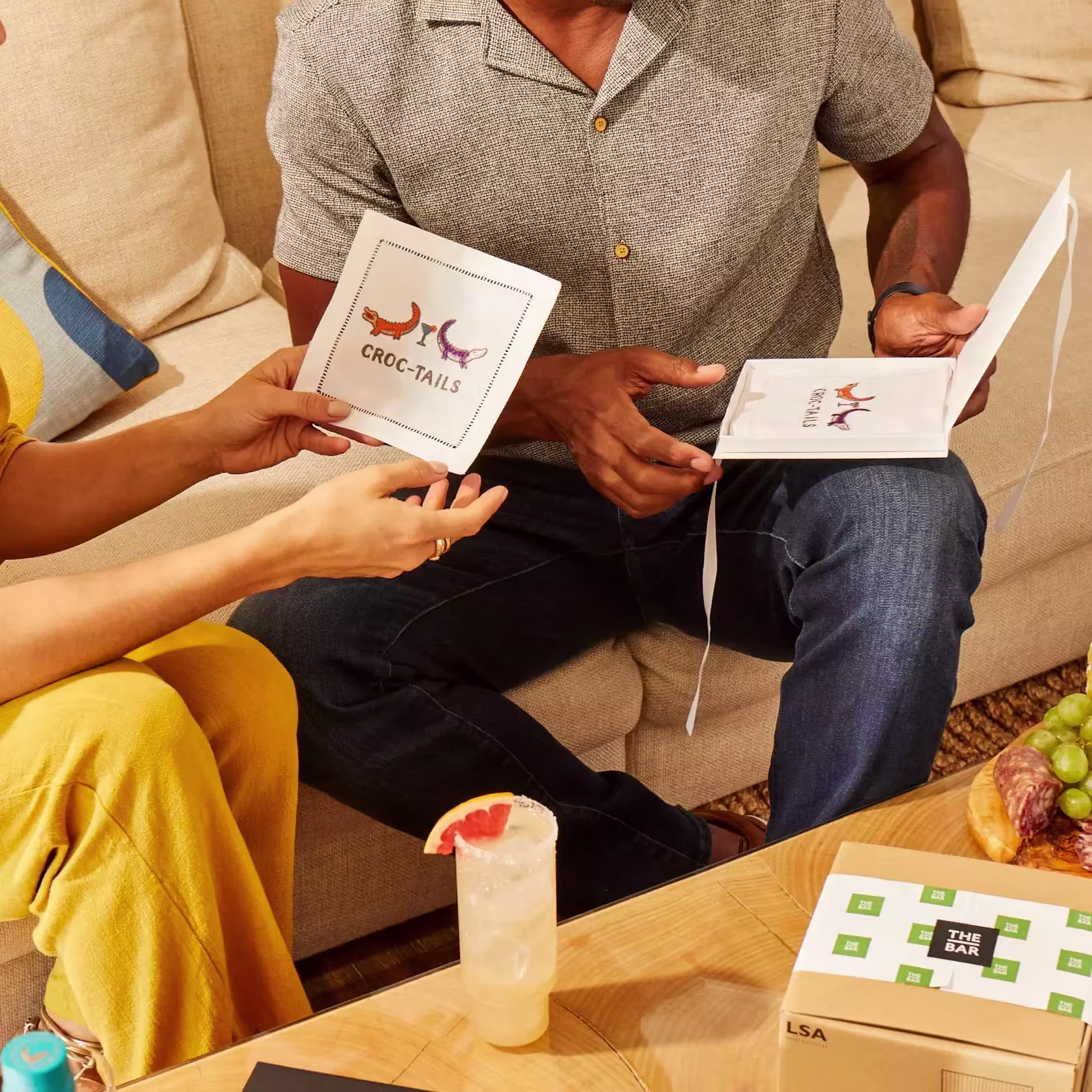 Two people sit on a couch holding cards featuring a crocodile and the word CROC-TAILS; a cocktail, grapes, and a box labeled THE BAR are on the table in front of them.