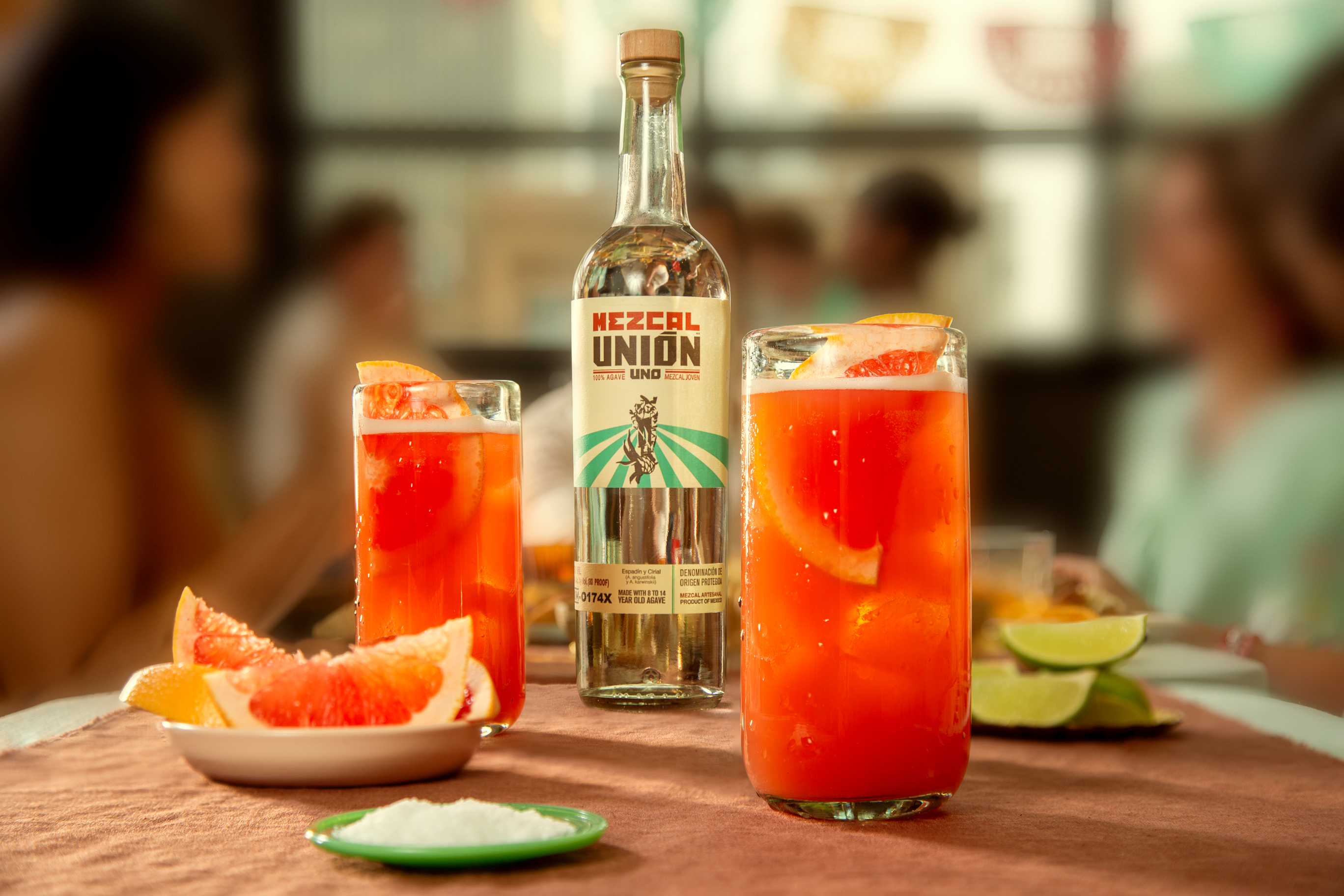 A bottle of Mezcal Unión is placed behind two glasses of red-orange cocktail with grapefruit slices, next to plates of grapefruit, lime wedges, and salt on a table.