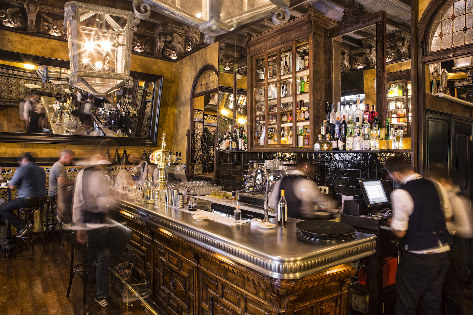 Muestra el interior del local Viva Madrid.Hay una larga barra de madera pulida con varias botellas de alcohol en estantes contra una pared con espejos. El bar tiene un aire sofisticado, con elaborados trabajos en madera y elementos decorativos.