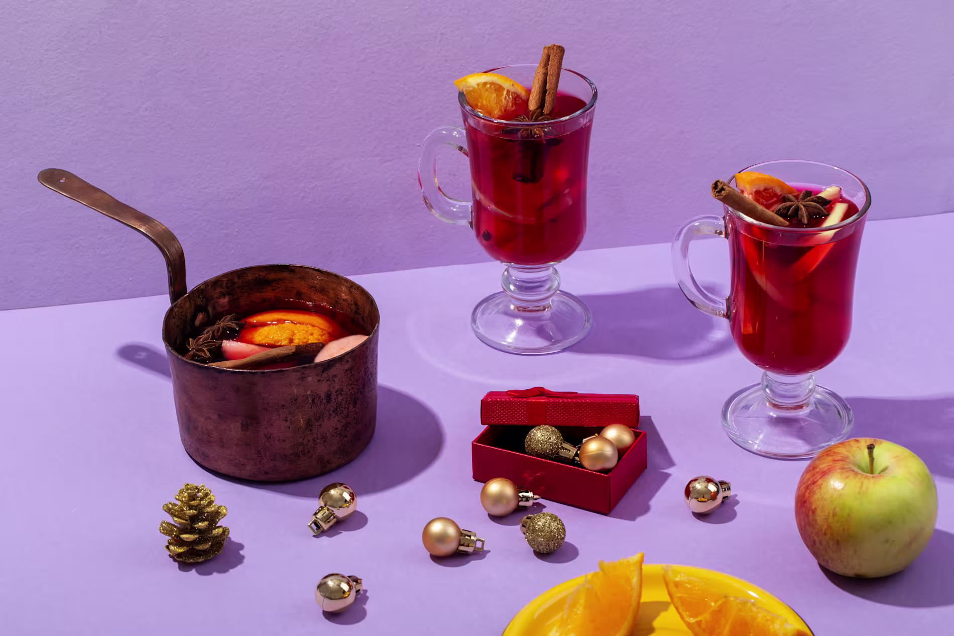 Two glass mugs and a copper pot of mulled wine with orange slices and spices, surrounded by a golden pinecone, apple, ornaments, and orange pieces on a purple surface.