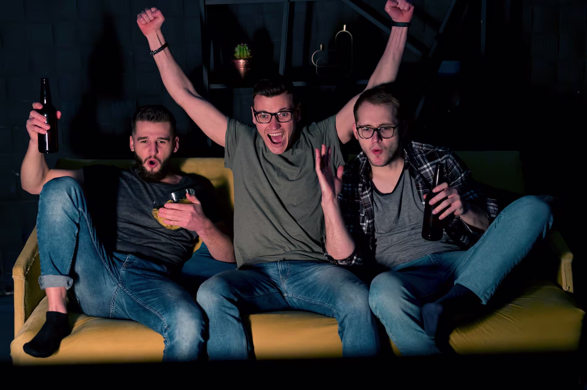 Three men sit on a yellow couch cheering and holding drinks while watching something, appearing excited and engaged.