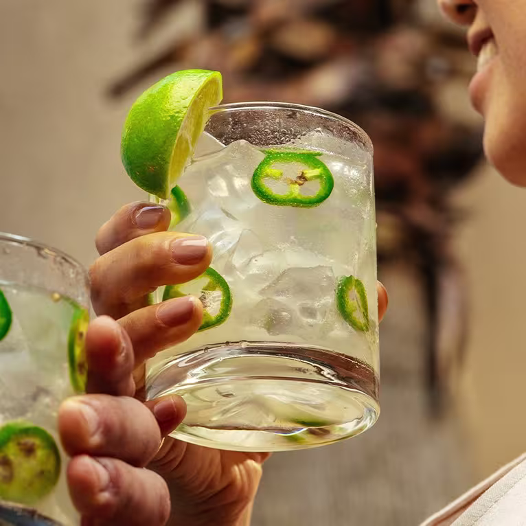 Două persoane țin margarita picantă cu garnitură de lime și felii de chili.