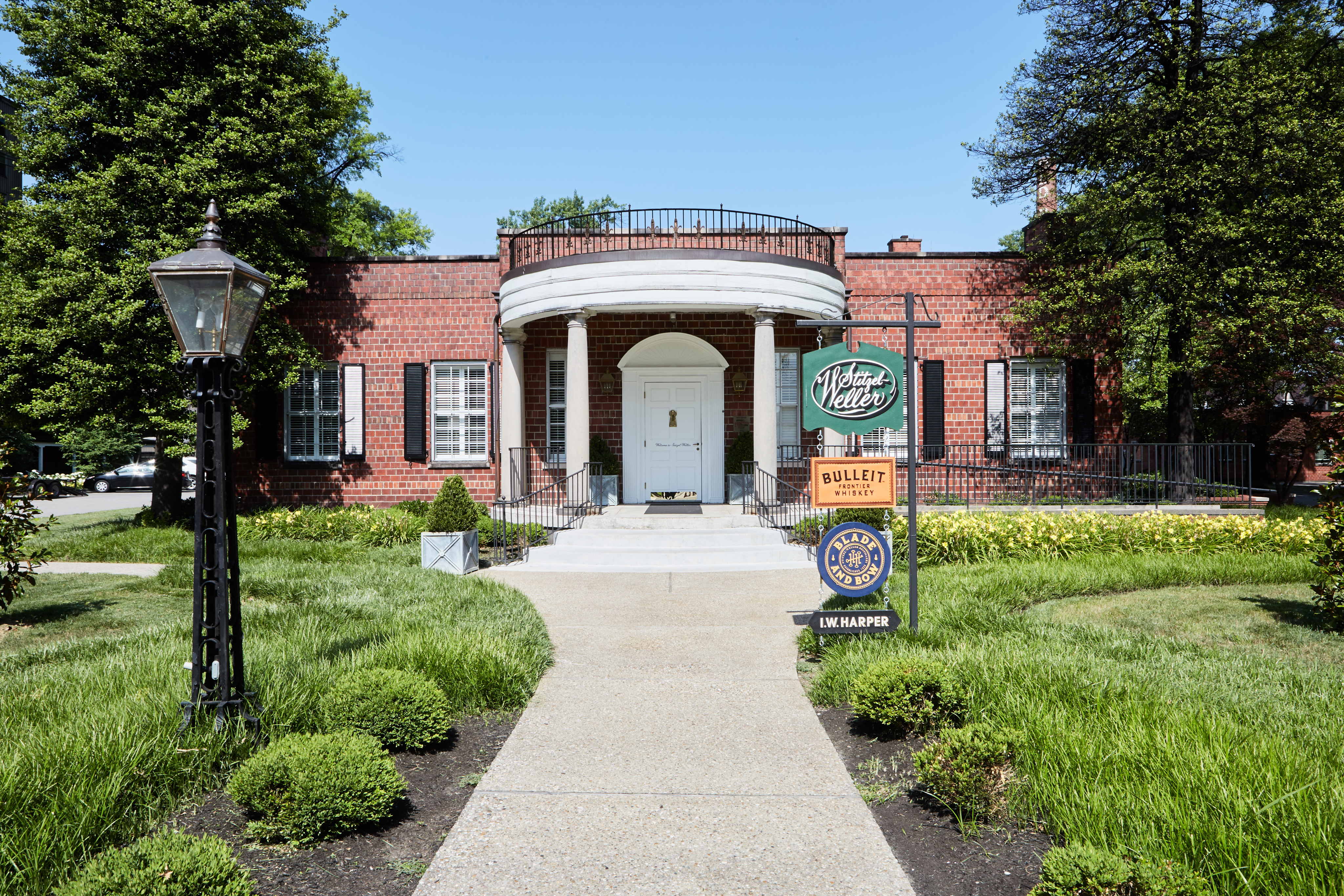 A red brick building with white columns and a central door, surrounded by greenery, with several whiskey brand signs displayed on the front lawn.