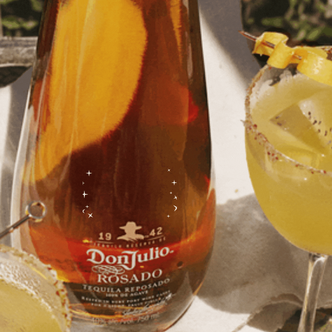 A bottle of Don Julio Rosado tequila sits beside two yellow cocktails in salt-rimmed glasses, garnished with lemon twists.