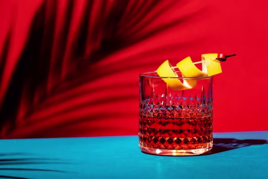 A cocktail with an orange peel garnish in a textured glass sits on a blue surface against a red background with a palm leaf shadow.